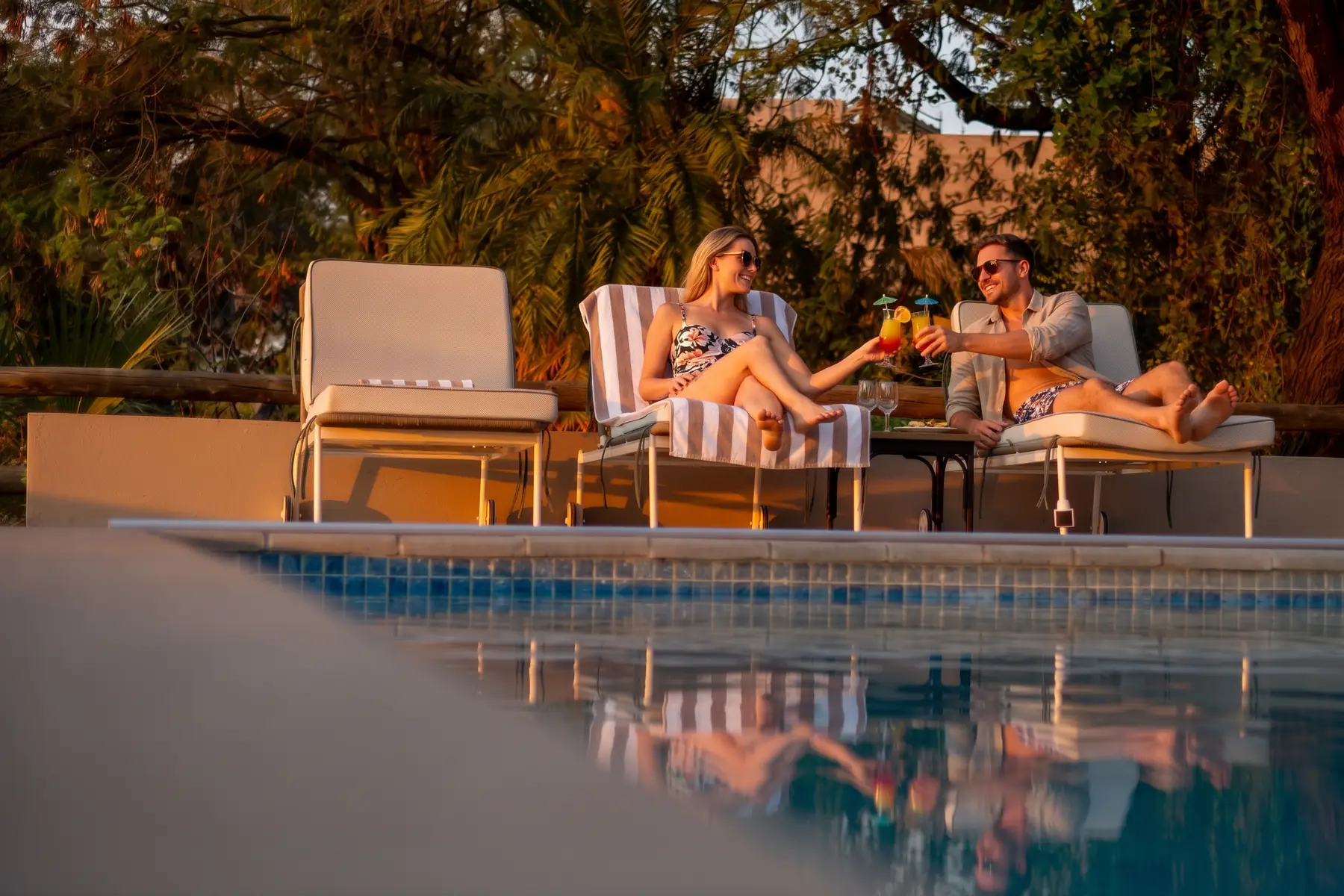 A couple toast with cocktails on loungers beside the pool at golden hour