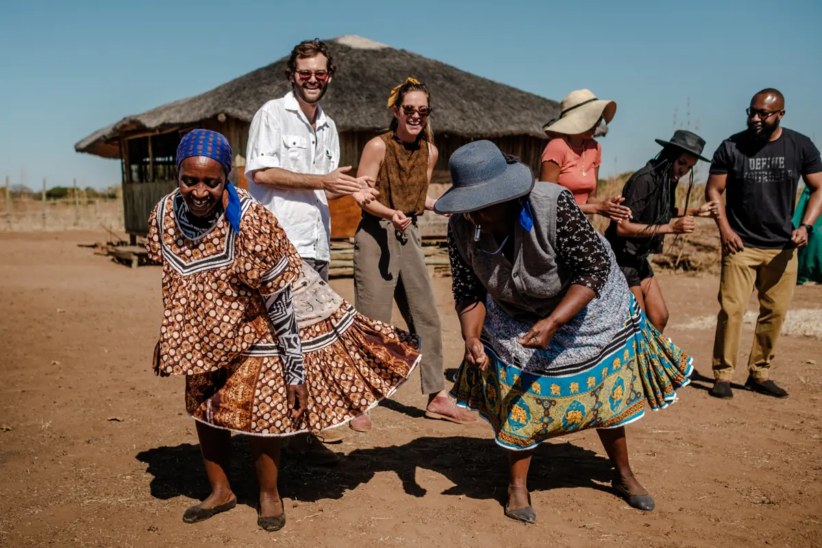 Guests dancing with women from the village outside a thatched hut
